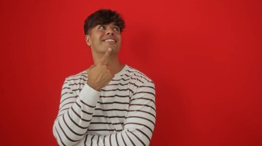 Yakışıklı, çizgili tişörtlü genç bir adam canlı kırmızı bir arkaplanda izole edilmiş, düşünceli bir şekilde yukarıya bakıyor..