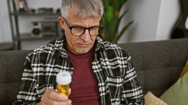 Evde gözlüklü, reçeteli ilaç kutusu okuyan olgun bir adam..
