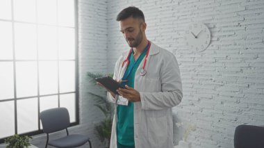 Beyaz önlük ve steteskop giymiş, parlak ışıklandırılmış bir kliniğin bekleme odasında elinde tablet tutan genç İspanyol erkek doktor, ekrana odaklanırken ciddi bir ifade sergiliyor..