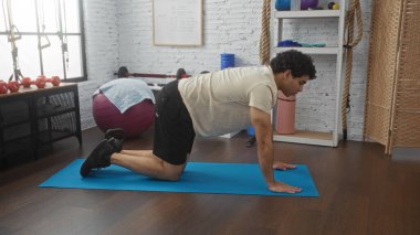 Genç İspanyol bir adam spor salonunda mavi bir paspasın üzerinde egzersiz yapıyor. Ekipmanlı bir spor salonu ve huzurlu bir atmosferi var..