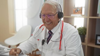 Kırmızı steteskoplu beyaz önlüklü yaşlı gri saçlı adam gülümsüyor ve iyi aydınlatılmış bir hastane kliniğinde kulaklık takıyor.