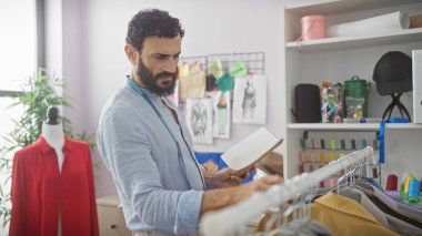 Modern terzi atölyesinde kumaşı inceleyen sakallı adam, renkli iplikler ve moda çizimleriyle çevrili..