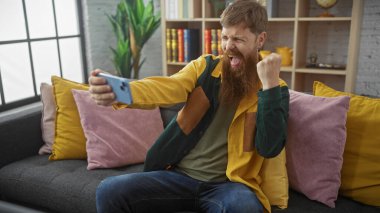 Heyecanlı sakallı, kızıl saçlı adam akıllı telefondan zaferi kutluyor içeride oturuyor ve gülümsüyor..