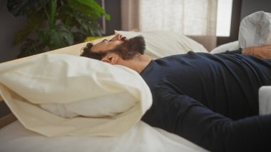 Sakallı adam yatak odasında huzur içinde dinleniyor. Beyaz yatak örtüsü ve yumuşak doğal ışıkla..