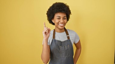Önlüklü neşeli Afrikalı Amerikalı kadın sarı arka planda bir fikir jesti ile poz veriyor..