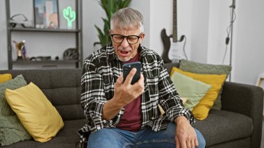 Olgun bir adam modern bir oturma odasında akıllı telefonuna tepki verir ve gözlük takarak şaşkınlığını ifade eder..