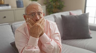 Gri saçlı, yaşlı, Kafkas görünümlü bir adam oturma odasında şoke olmuş bir şekilde oturuyor, kapalı bir koltukta oturuyor ve elleri ağzını ve açık bakışlarını kapatıyor..