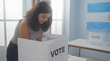 Latin kökenli genç bir esmer kadın, Arjantin 'de bir üniversite odasında oy veriyor. Görünür bir oy verme kabini ve bir Arjantin bayrağı ile kapalı bir yerde duruyor..