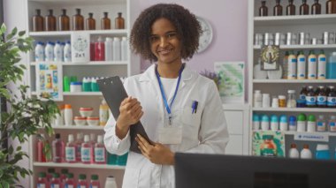 Afrika kökenli Amerikalı kadın eczacı kıvırcık saçlı ve elinde bir pano olan iyi stoklanmış bir eczanede gülümsüyor..
