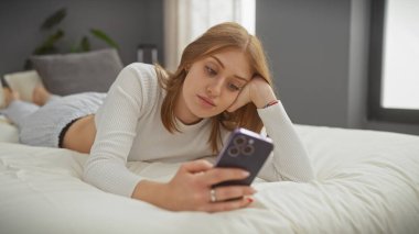 Beyaz bir kadın evde yatakta uzanıyor, kapalı bir ortamda akıllı telefonuna bakıyor..
