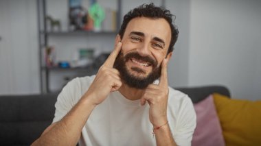 Bearded man smiles indoors touching face in cozy home setting clipart