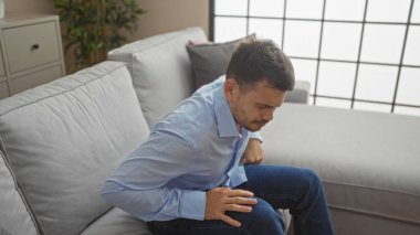 Sakallı genç bir İspanyol, gri bir kanepede oturuyor şık bir oturma odasında, sırt ağrısı yüzünden acı çekiyor gibi görünüyor..