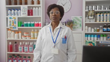 Eczanede duran gözlüklü ve beyaz önlüklü kadın eczacı duvardaki görünür saatin altında çeşitli sağlık ürünleriyle dolu raflarla çevrili.