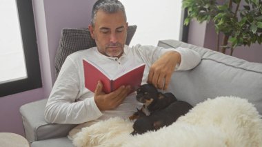 Oturma odasındaki kanepede kitap okuyan orta yaşlı bir adam yanında iki Chihuahua ile birlikte, ev ortamında rahat bir ortam yansıtıyor..