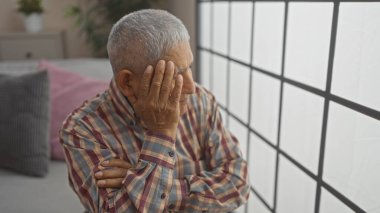 Evde gri saçlı, olgun İspanyol bir adam. Oturma odasındaki pencereden dışarı bakıyor. Ekose bir gömlekle elini uzatırken kollarını çaprazlıyor..