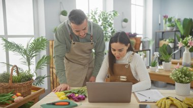 Önlüklü bir adam ve kadın, çiçekçi dükkanında dizüstü bilgisayar kullanıyor. Etrafı çiçeklerle çevrili, küçük bir işletmedeki takım çalışmasını temsil ediyor..