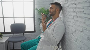 Hastanenin bekleme odasında sakallı ve steteskoplu yakışıklı genç adam akıllı telefondan sesli mesaj gönderiyor.