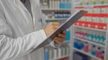Arkaplanda ilaç rafları olan bir eczaneye notlar yazan beyaz önlüklü bir erkek eczacı.