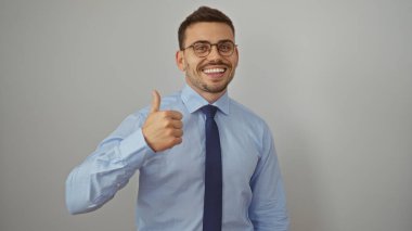 Yakışıklı, sakallı ve gözlüklü genç bir adam beyaz bir arka planda güvenle gülümserken başparmağını kaldırıyor..