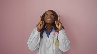 Güzel, genç, Afro-Amerikalı, kıvırcık saçlı bir bayan doktor neşeli bir şekilde gülümsüyor ve pembe arka planda dikilirken yukarı doğru işaret ediyor..