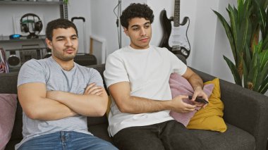 İki adam kapalı bir kanepede oturuyor, biri uzaktan kumandalı, rahat ve rahat bir atmosfer yayıyor..