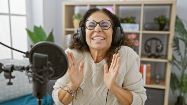 Profesyonel mikrofon ve kulaklıkla modern bir radyo stüdyosunda kayıt yaparken gülümseyen İspanyol kadın..