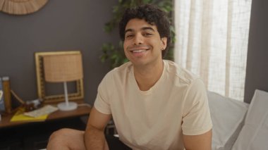 Yakışıklı, genç, İspanyol bir adam sıcak, iyi dekore edilmiş bir yatak odasında yatağında oturmuş evinde sıcak ve davetkar bir sahne yaratırken gülümsüyor..