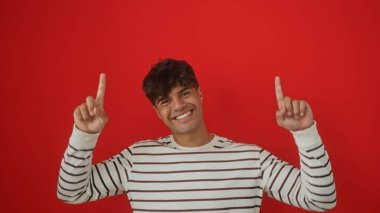 İzole edilmiş kırmızı bir arka planda çizgili süveter içinde gülümseyen yakışıklı İspanyol bir adam, iki eliyle yukarıya bakıyor..