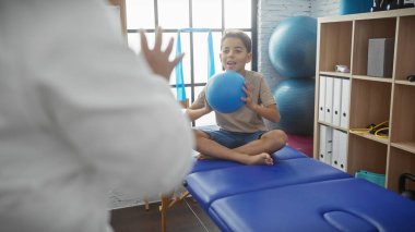 Fizyoterapi seansındaki bir çocuk, klinikteki bir terapistin rehberliğinde mavi topuyla oynar.