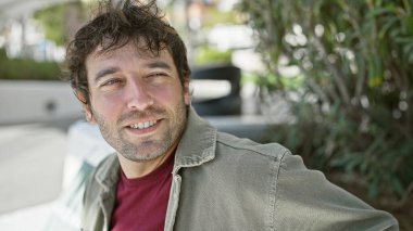 Sakallı yakışıklı, genç, İspanyol bir adam zengin bir şehir parkında kibarca gülümsüyor..