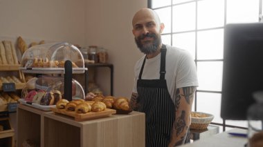 Bir pastanede yakışıklı İspanyol bir adam cam bir pasta vitrininin yanında duruyor, ev ortamında mutlu bir şekilde gülümsüyor.