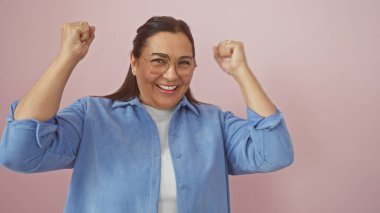 Neşeli, orta yaşlı İspanyol bir kadın pembe bir arka plana karşı yumruklarını kaldırarak kutluyor..