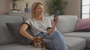 Gülümseyen sarışın bir kadın oturma odasında dinleniyor. Evde akıllı telefonuna bakarken köpeğini okşuyor..