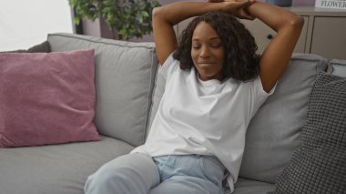 Beyaz t-shirt ve kot pantolon giyen kıvırcık saçlı Afro-Amerikalı bir kadın, bir ev ortamında yakalanmış..