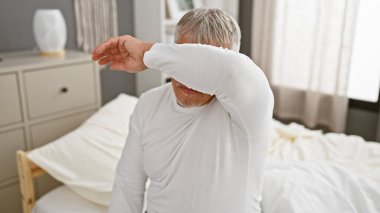 Mature grey-haired man covers face with arm in bedroom setting, embodying distress or morning fatigue. clipart