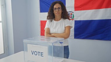 Dominik Cumhuriyeti 'nde seçmen odasında kollarını kavuşturmuş bir İspanyol kadın. Arka planda şeffaf bir oy sandığının ve bayrağın yanında duruyor..