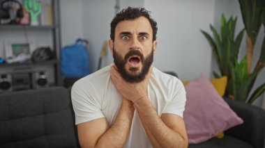 Modern bir oturma odasında beyaz gömlekli, şok olmuş sakallı bir adam boynunu sıkıyor..