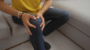 Orta yaşlı İspanyol bir adam evdeki bir kanepede oturuyor, dizini rahatsızlıkla tutuyor, sarı gömlek ve kot da dahil olmak üzere günlük kıyafetler giyiyor, elleri ve dizlerini detaylı bir şekilde gösteriyor.