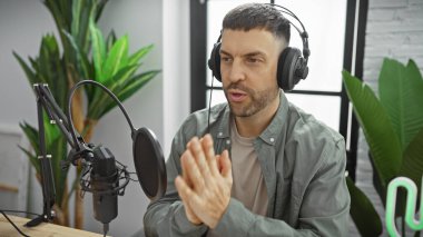 Handsome hispanic man with a beard talking into a microphone in a podcast studio clipart