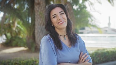 Orta yaşlı, gülümseyen İspanyol bir kadın. Kolları çaprazlanmış. Açık havada, yemyeşil bir parkta duruyor.