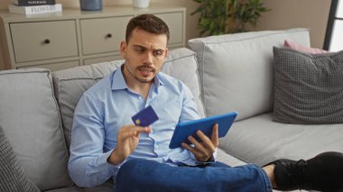 Sakallı, yakışıklı bir İspanyol adam oturma odasında elinde bir kredi kartı ve tablet ile online bir aktiviteye girişiyor..