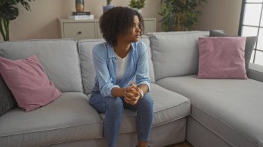 Kıvırcık saçlı genç siyah bir kadın şık bir oturma odasında gri bir kanepede oturuyor açık mavi bir gömlek ve mavi kot pantolon giyiyor, dalgın dalgın bakıyor..