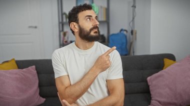 Düşünceli sakallı beyaz gömlekli adam, modern oturma odasındaki gri koltukta otururken...