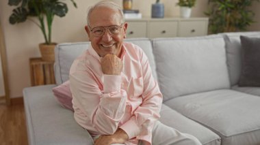 Yakışıklı, orta yaşlı, beyaz saçlı, rahat bir oturma odasında gülümseyen, modern bir iç mekan ve iç mekan bitkisi olan beyaz bir adam..