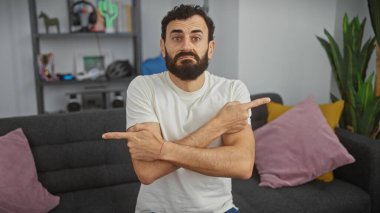 Sakallı adam modern bir oturma odasında çapraz kollarla el kol hareketi yapıyor..