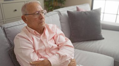 Yaşlı, gri saçlı, düşünceli bir ifadeyle oturma odasında oturan, yumuşak ışıklandırma ve modern mobilyalarla rahat bir iç mekan ortamı sergiliyor..