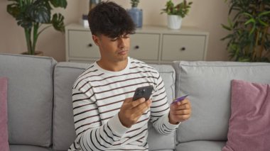 Modern bir oturma odasında bitki ve dekorasyonlarla dolu rahat bir koltukta otururken elinde kredi kartı ve akıllı telefon kullanan yakışıklı İspanyol bir adam..