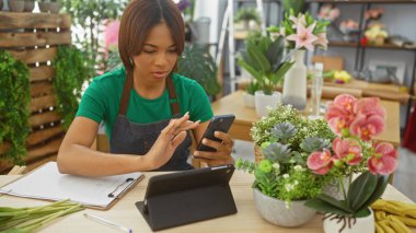 Çiçekçi dükkanında akıllı telefon kullanan Afrikalı Amerikalı kadın. Etrafı buketler ve bitkilerle çevrili..