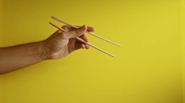 Sarı arka planda çubuk tutan bir adam minimalist bir ortamda geleneksel yemek aletlerini sergiliyor.