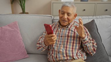 Yaşlı İspanyol bir adam oturma odasında rahat bir kanepede otururken akıllı telefonuyla video konferansı yapıyor..